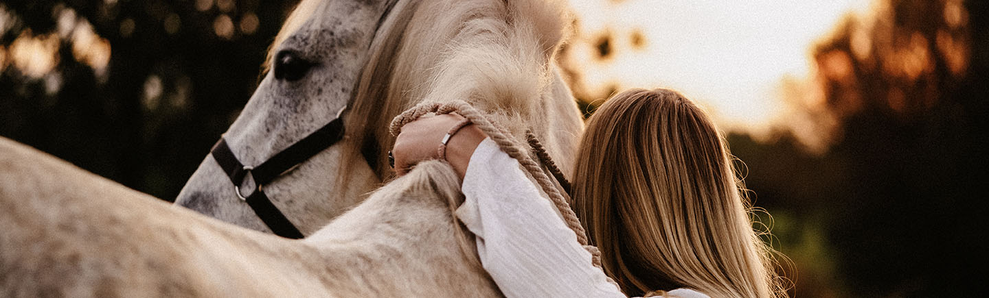 Portrait of a blond young woman from behind embracing her white horse in nature